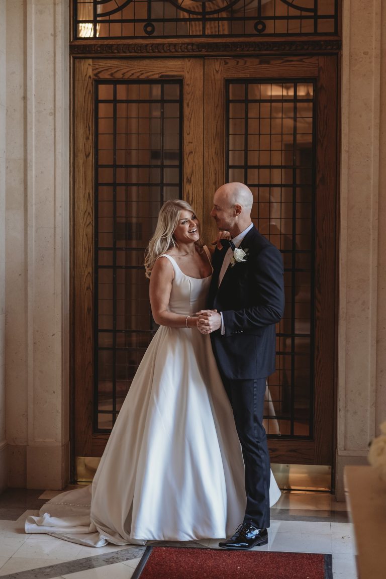 Bride and groom share a romantic moment smiling at each other during their first dance inside a luxurious London wedding venue, standing by classic wooden doors with ornate glass panels.