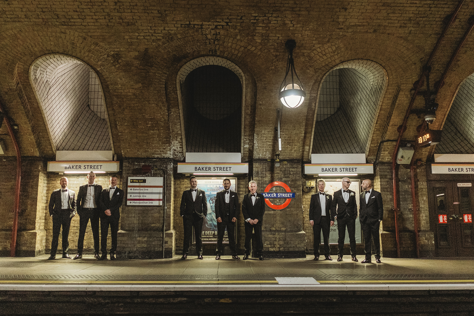 Groom and groomsmen dressed in black-tie attire standing under the iconic arched brick ceiling at Baker Street Underground Station in London, striking a cinematic pose before the wedding.