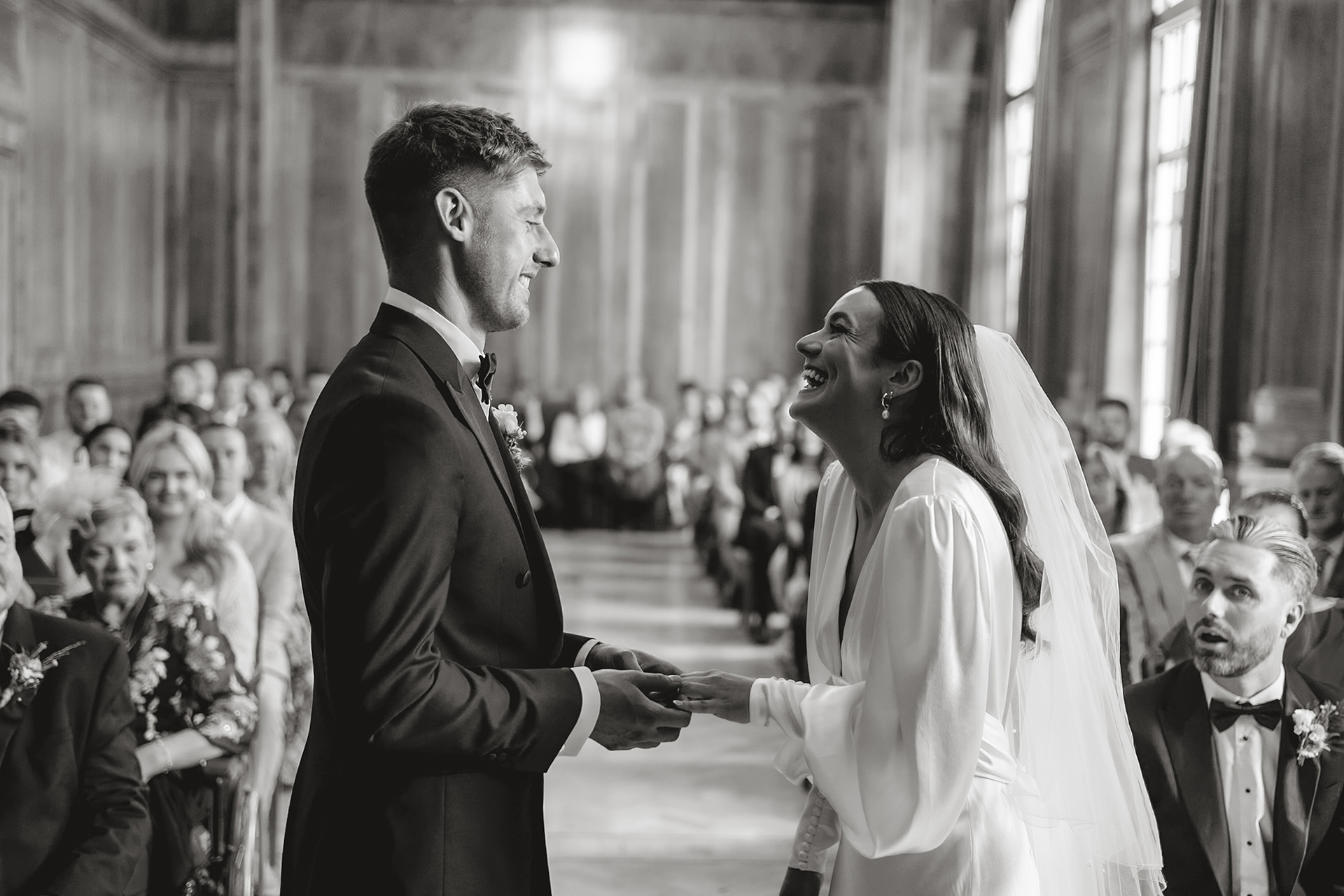 The bride and groom share a joyful moment during their wedding ceremony inside the wood-panelled Council Chamber at Hackney Town Hall. The groom places the ring on the bride’s finger as she laughs, surrounded by smiling guests in the background.