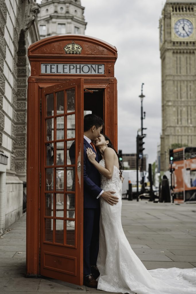 A bride and groom share a romantic embrace inside a classic red London telephone box, with Big Ben in the background. The bride wears a white lace gown with a long train, while the groom is in a navy suit and red tie. Captured as part of a London pre-wedding shoot before their Singapore wedding.