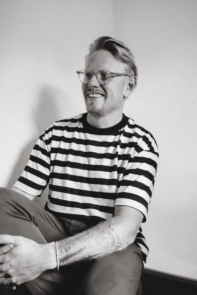 Black and white portrait of Jon, a professional wedding photographer in London, smiling in a striped t-shirt. Relaxed lifestyle photography style showcasing personality and creativity.