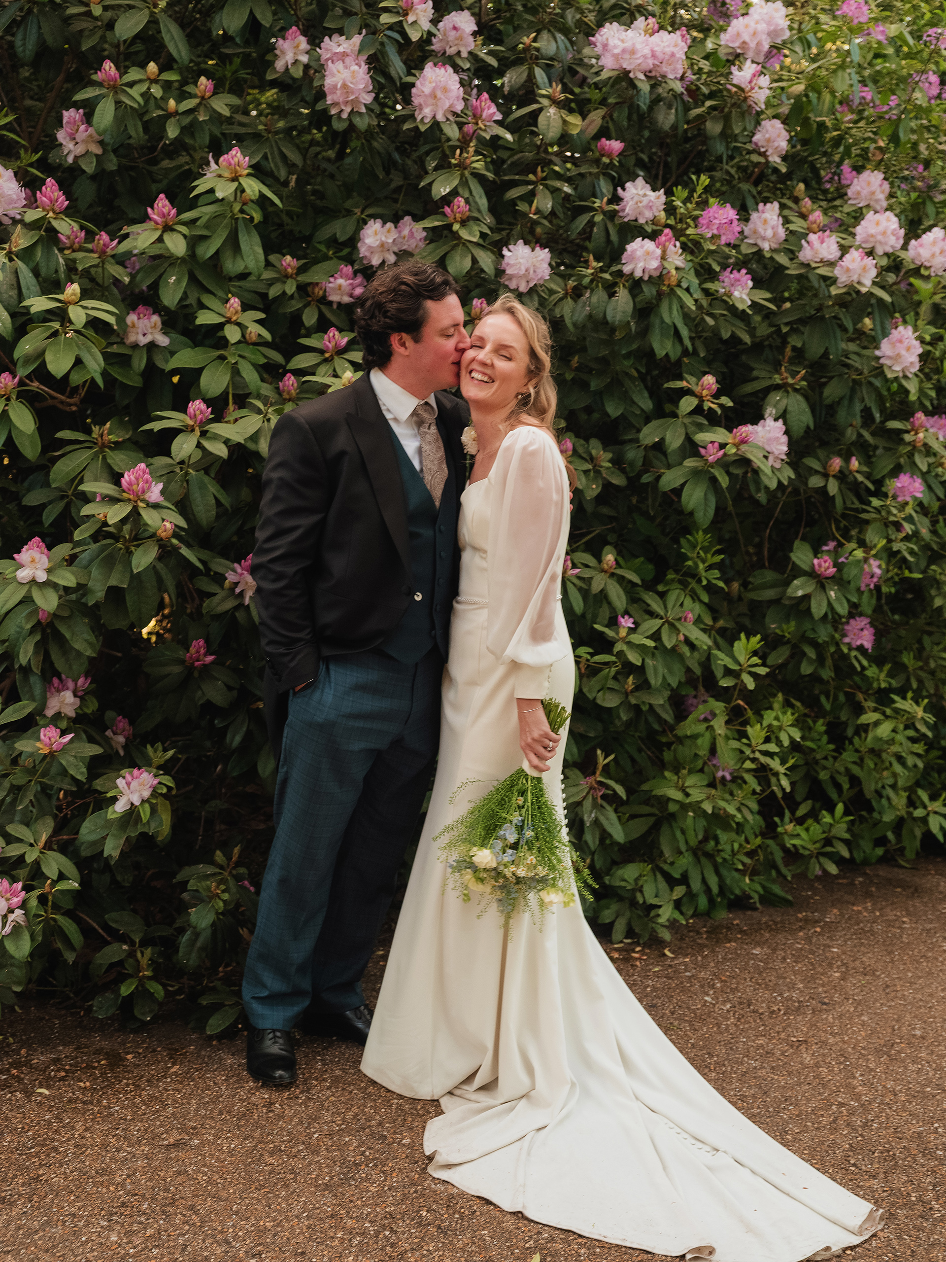 Groom kisses the bride on the cheek as they stand in front of blooming pink flowers, both smiling warmly.