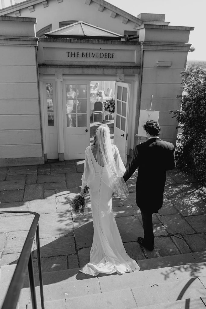 Black-and-white photo of the bride and groom walking hand in hand down stone steps toward the reception venue entrance.